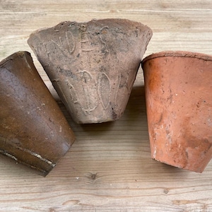 May include: Three weathered terracotta plant pots. The pots are conical, with colours ranging from light brown to reddish-brown, showing texture and imperfections. They are arranged on a wooden surface.