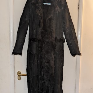 Può includere: Un cappotto lungo nero con collo e finiture in pelliccia, appeso a una gruccia bianca. Il cappotto sembra essere realizzato in pelle o camoscio, con un design patchwork. Il cappotto ha maniche lunghe e due tasche. Il cappotto è appeso a una porta bianca.