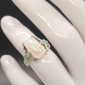 May include: A silver ring with a large, rectangular, iridescent opal stone set in a silver setting. The ring is accented with small, green stones.