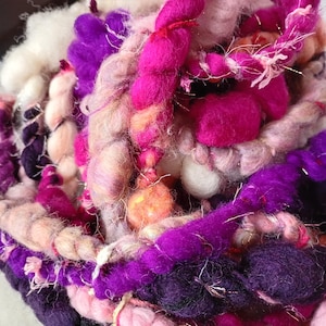 Op de afbeelding: Close-up van een levendige, getextureerde garenbundel. Het garen heeft een mix van kleuren, waaronder tinten paars, roze en wit. De vezels variëren in dikte en textuur, wat een visueel interessant en tactiel effect creëert. Ideaal voor handwerkprojecten.