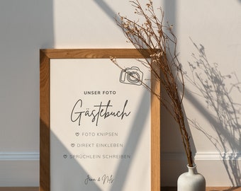 May include: A framed print with the words "UNSER FOTO GÄSTEBUCH" in elegant script. The print includes a line drawing of a camera and instructions. The frame is made of light brown wood. A small white vase with dried flowers is next to the frame.