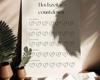 May include: A beige wedding countdown poster with the text "Hochzeits-countdown." The poster features rows of heart-shaped outlines, each with a number inside, counting down the weeks until the wedding. The poster is displayed on a wooden surface.