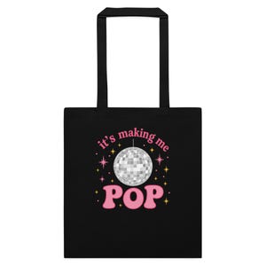 Może przedstawiać: Czarna torba typu tote z białą grafiką kuli dyskotekowej i różowym napisem "it's making me POP". Torba ma czarne uchwyty i jest przeznaczona do przenoszenia przedmiotów.