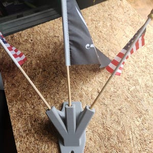 Puede incluir: Un soporte de pared gris impreso en 3D con tres ranuras para mástiles de bandera. El soporte tiene un agujero para colgar. Tres mástiles de bandera se insertan en las ranuras, cada uno sosteniendo una bandera. Las banderas son una bandera estadounidense, una bandera negra con una luna creciente blanca y una estrella, y una bandera blanca con una franja roja.