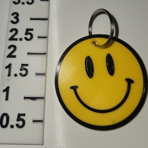 May include: Yellow smiley face keychain with a black outline. The keychain is attached to a silver ring.