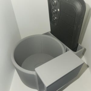 May include: A gray 3D-printed car cup holder with an integrated phone holder. A black smartphone is placed in the phone holder. The cup holder has a cylindrical shape and a rectangular side extension.