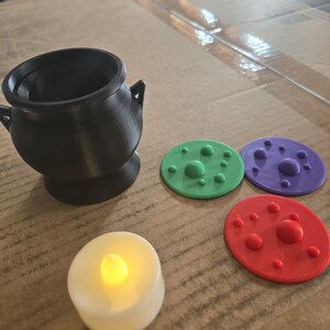 May include: A black cauldron with two side handles, a white battery-operated tea light with a glowing yellow flame, and three flat circular discs in green, purple, and red, each featuring raised spherical bumps on the top surface.