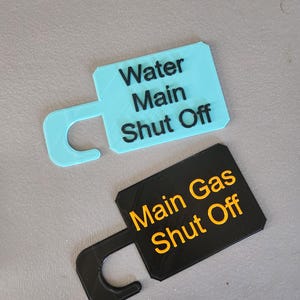 May include: Two rectangular, 3D-printed tags with a curved handle. One is light blue with the text "Water Main Shut Off" in black. The other is black with the text "Main Gas Shut Off" in orange. Both are on a gray surface.