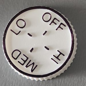 May include: A white control knob with a ridged edge and the words "LO", "OFF", "MED", and "HI" in dark purple. The knob has a series of dashes indicating settings. The background is a solid gray.