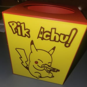 May include: A yellow tissue box with a red interior and top rim. The box features a cartoon Pikachu character with the text "Pik Achu!" in brown lettering. The design is a parody of the popular Pokemon character.