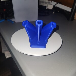 Könnte beinhalten: Ein leuchtend blaues, 3D-gedrucktes Objekt mit einem einzigartigen, fächerartigen Design. Es hat drei abgewinkelte Arme, jeder mit einer kreisförmigen Öffnung am Ende, die mit einer zentralen vertikalen Stütze verbunden sind. Das Objekt steht auf einer weißen, kreisförmigen Basis.
