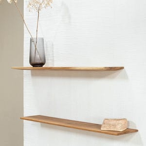 Op de afbeelding: Twee zwevende houten planken aan een witte muur. De bovenste plank bevat een grijze glazen vaas met gedroogde bloemen. De onderste plank heeft een kleine, lichtgekleurde steen. De planken zijn van lichtgekleurd hout.
