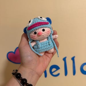 May include: A handmade crochet piglet doll wearing a blue hat with white eyes and pink accents, and a matching blue outfit. The doll has a pink snout and cheeks. The doll is held in a hand.
