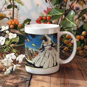 Puede incluir: Taza de café de cerámica blanca con un gráfico de una criatura parecida a Godzilla que lleva un vestido de novia blanco y está de pie frente a un horizonte urbano. La taza tiene un asa negra.