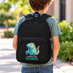 May include: A black backpack featuring a graphic of a light blue iguana with orange spikes. Below the iguana, the text reads "I will do what IGUANA" in white and teal lettering. The bag has a front zippered pocket and black shoulder straps.