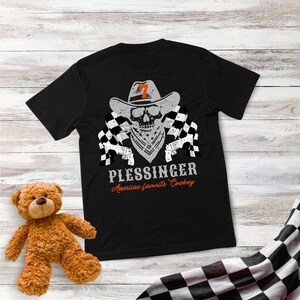 May include: Black t-shirt featuring a skull wearing a cowboy hat with the number 7, a bandana, and checkered flags. The text "PLESSINGER Americas favorite Cowboy" is printed below the graphic.