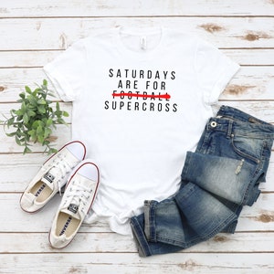 Peut inclure: T-shirt blanc avec un texte noir qui dit "Saturdays are for FOOTBALL SUPERCROSS" avec le mot "FOOTBALL" barré en rouge.