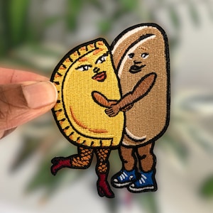 Könnte beinhalten: Gestickter Aufnäher mit einer gelben Empanada und einem braunen Hotdog, die sich umarmen. Die Empanada hat ein Gesicht mit rotem Lippenstift und der Hotdog hat ein braunes Gesicht. Beide Figuren tragen Schuhe.