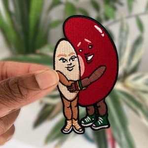 Puede incluir: Parche bordado con dos personajes de dibujos animados, una judía roja y una almendra tostada, abrazándose. La judía tiene una cara sonriente y la almendra tiene una expresión de sorpresa. Ambos personajes llevan zapatos y tienen brazos y piernas.