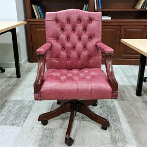Peut inclure: Un fauteuil de bureau en cuir bordeaux avec un dossier et une assise capitonnés. La chaise a des accoudoirs en bois et un piètement à cinq branches avec des roulettes. La chaise est dans une pièce avec une bibliothèque en bois et un bureau.
