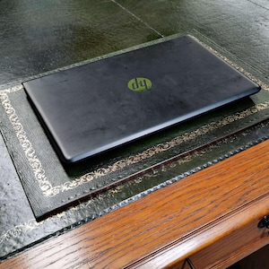 May include: A black laptop computer with the HP logo on the lid is sitting on a green leather desk pad with gold trim. The desk pad is on a wooden desk.