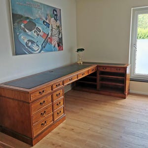 Può includere: Una grande scrivania in legno a forma di L con piano in pelle nera. La scrivania ha più cassetti e ripiani ed è realizzata in legno dai toni caldi. Un poster di auto da corsa vintage è appeso al muro sopra la scrivania.