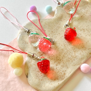 Puede incluir: Cuatro abalorios de fresa de vidrio rojo y transparente en cuerdas. Los abalorios cuelgan de un plato de cerámica blanco con un glaseado marrón moteado. El plato está sobre una superficie rosa con pompones amarillos.