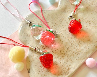 May include: Four red and clear glass strawberry charms on strings. The charms are hanging from a white ceramic plate with a speckled brown glaze. The plate is on a pink surface with yellow pom-poms.