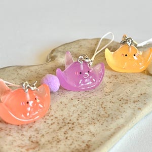 May include: Three colorful, translucent, cartoon-like stingray charms with happy faces. The charms are attached to phone straps with silver rings and beads.