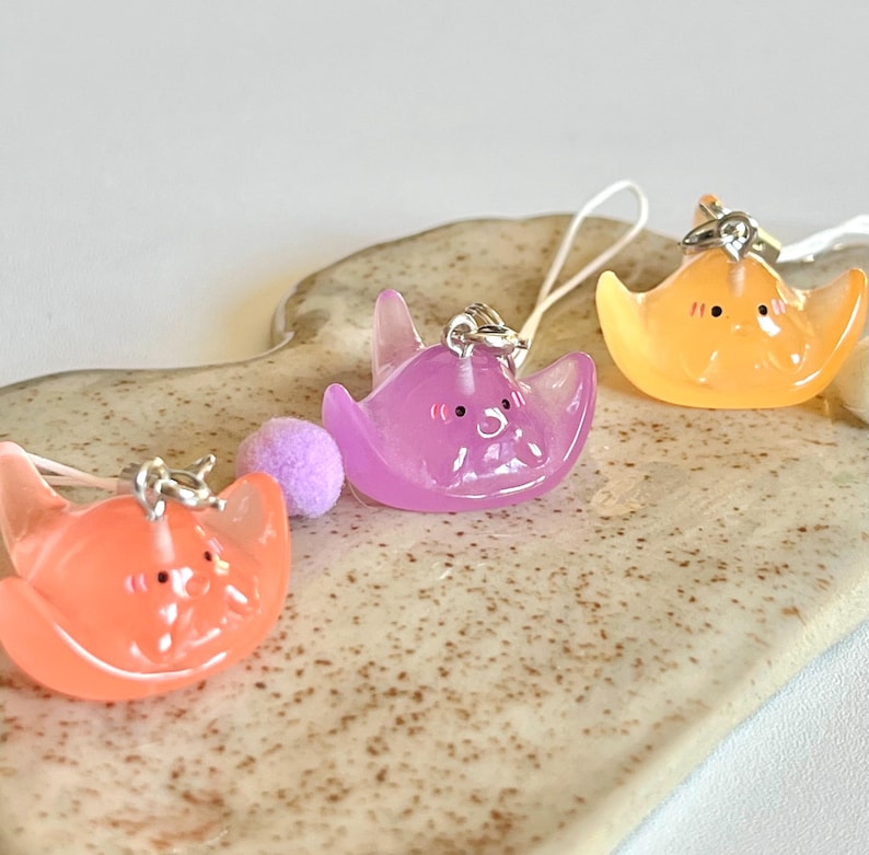 May include: Three colorful, translucent, cartoon-like stingray charms with happy faces. The charms are attached to phone straps with silver rings and beads.