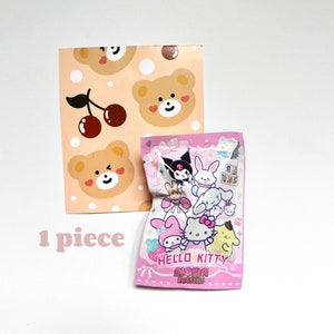 May include: A pink and white Hello Kitty eraser pack featuring a variety of Hello Kitty characters. The pack is on a white background with a pink and brown patterned paper behind it. The text on the pack reads "Hello Kitty Erasers".