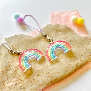 Puede incluir: Dos correas de teléfono con un encanto de arcoíris y un cierre plateado. Los encantos están hechos de resina transparente con un efecto de brillo de arcoíris. Las correas son moradas y blancas.