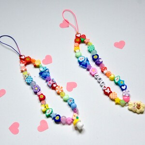Op de afbeelding: Twee kleurrijke kralen telefooncharms met regenboogkleuren en verschillende bedels, waaronder harten, sterren, bloemen en smileygezichten. Een bedel heeft het woord "JOY" gespeld in kralen en de andere heeft een kleine paddenstoelbedel.