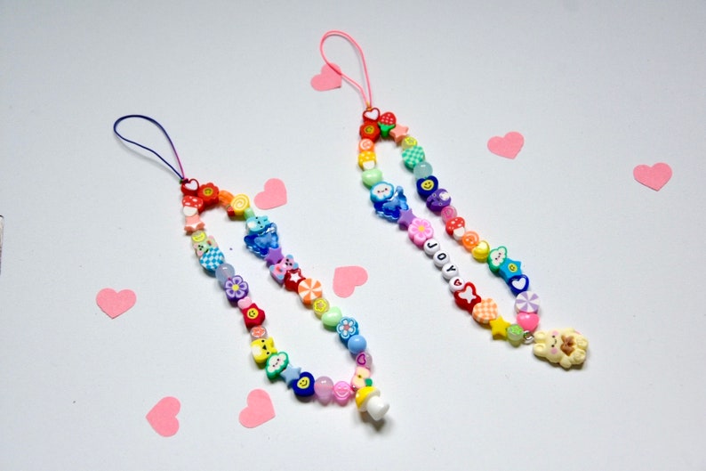 Op de afbeelding: Twee kleurrijke kralen telefooncharms met regenboogkleuren en verschillende bedels, waaronder harten, sterren, bloemen en smileygezichten. Een bedel heeft het woord "JOY" gespeld in kralen en de andere heeft een kleine paddenstoelbedel.