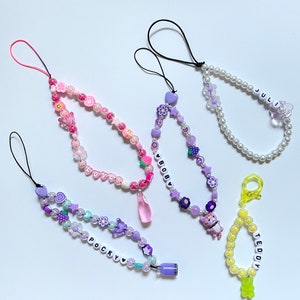 Op de afbeelding: Vier kleurrijke kralen telefooncharms met verschillende namen en ontwerpen. De charms zijn gemaakt met kleine kralen in verschillende kleuren, waaronder roze, paars, wit en geel. De charms hebben kleine bedels, zoals bloemen, sterren en harten. De namen op de charms zijn "LOVE", "BOBBY", "POCKY" en "TEDDY".