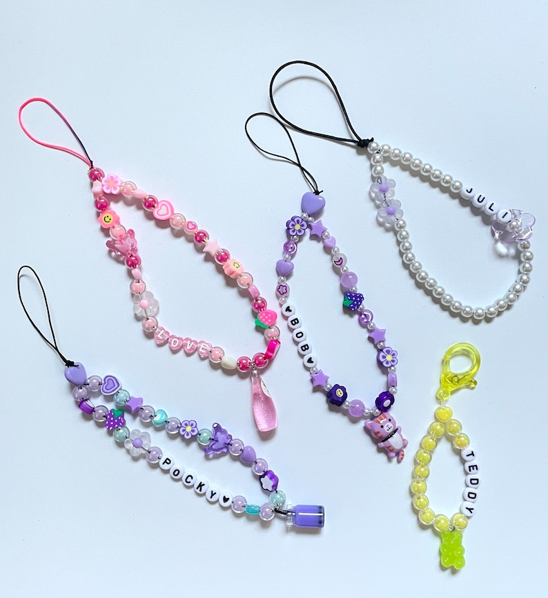 Op de afbeelding: Vier kleurrijke kralen telefooncharms met verschillende namen en ontwerpen. De charms zijn gemaakt met kleine kralen in verschillende kleuren, waaronder roze, paars, wit en geel. De charms hebben kleine bedels, zoals bloemen, sterren en harten. De namen op de charms zijn "LOVE", "BOBBY", "POCKY" en "TEDDY".