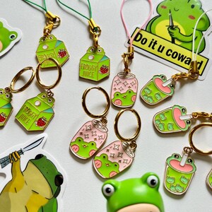 Può includere: Una collezione di portachiavi e adesivi a tema rana verdi e rosa. I portachiavi includono cartoni di "Froggy Juice", lattine di soda rosa e tazze di tè boba. Gli adesivi presentano illustrazioni di rane con il testo "Do it u coward".