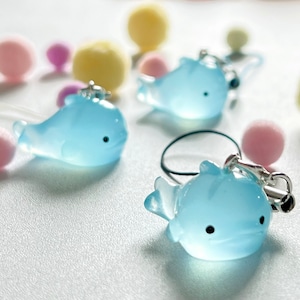 May include: Three light blue whale charms with black eyes. The whales are made of a translucent material and have a shiny finish. The charms are attached to a string or cord.