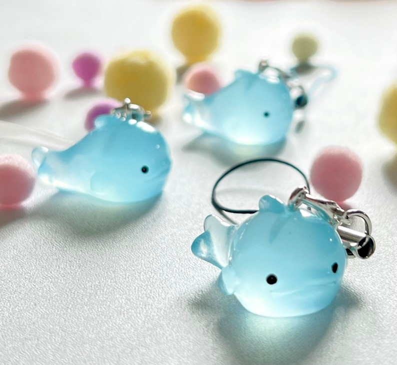 May include: Three light blue whale charms with black eyes. The whales are made of a translucent material and have a shiny finish. The charms are attached to a string or cord.