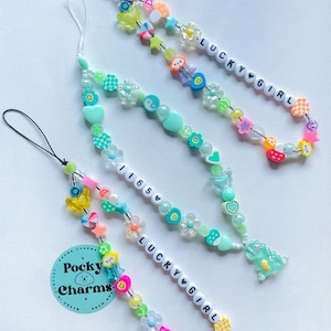 Op de afbeelding: Drie kleurrijke kralen telefooncharms met de tekst "Lucky Girl" en "1165" op witte kralen. De charms zijn gemaakt met verschillende kleuren kralen, waaronder groen, roze, geel en blauw. De charms hebben ook kleine charms, zoals harten, sterren en bloemen. De charms zijn bevestigd aan zwarte en witte telefoonbandjes.