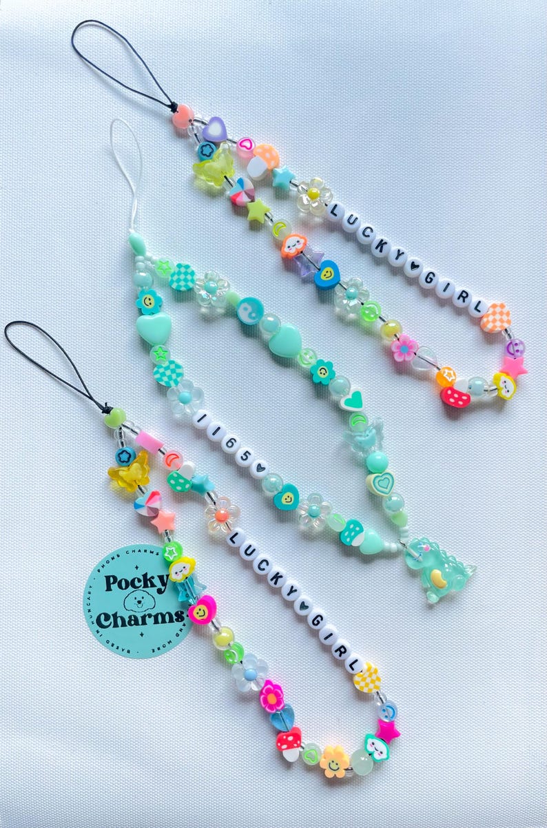 Op de afbeelding: Drie kleurrijke kralen telefooncharms met de tekst "Lucky Girl" en "1165" op witte kralen. De charms zijn gemaakt met verschillende kleuren kralen, waaronder groen, roze, geel en blauw. De charms hebben ook kleine charms, zoals harten, sterren en bloemen. De charms zijn bevestigd aan zwarte en witte telefoonbandjes.