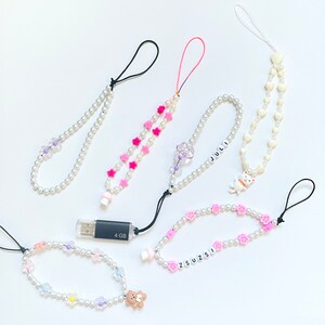 Op de afbeelding: Vijf kleurrijke telefooncharms gemaakt van witte parel kralen en verschillende bedels. De bedels zijn roze sterren, paarse bloemen, een witte paddenstoel, een witte kat en een witte beer. De charms zijn bevestigd aan zwarte en witte koorden.