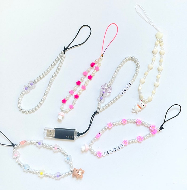 Op de afbeelding: Vijf kleurrijke telefooncharms gemaakt van witte parel kralen en verschillende bedels. De bedels zijn roze sterren, paarse bloemen, een witte paddenstoel, een witte kat en een witte beer. De charms zijn bevestigd aan zwarte en witte koorden.