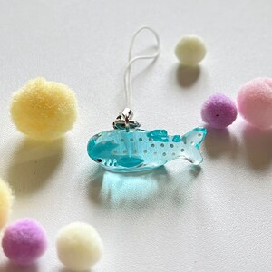 May include: A light blue whale shark charm with white spots, attached to a white string phone charm.