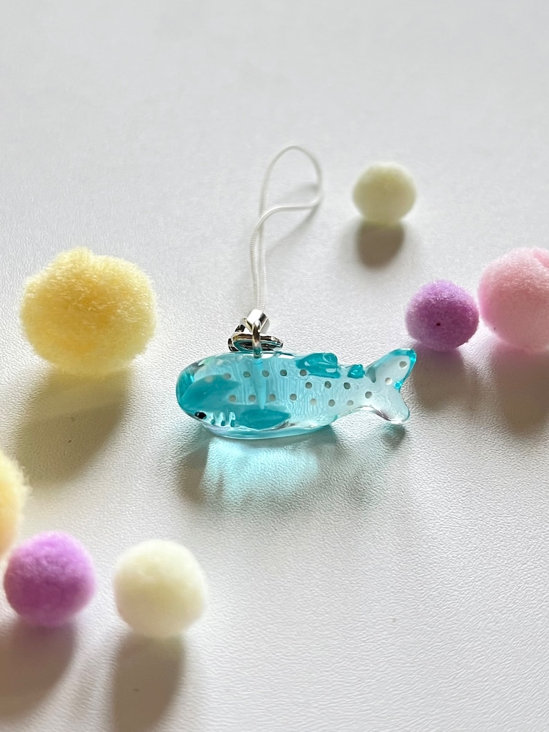May include: A light blue whale shark charm with white spots, attached to a white string phone charm.