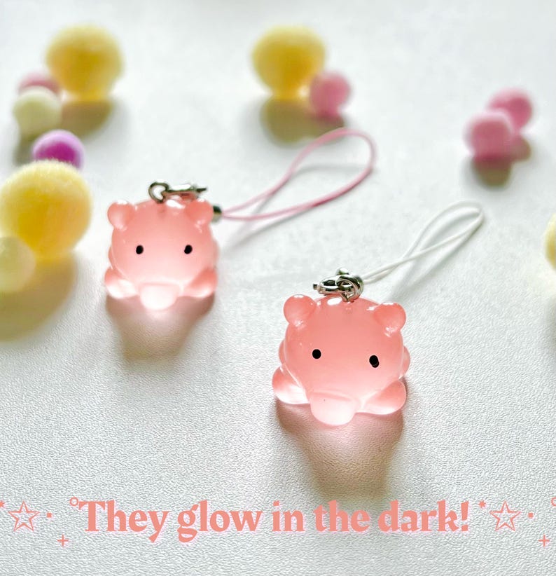 May include: Two pink glow-in-the-dark charms shaped like a cartoon animal with black eyes and a small mouth. The charms are attached to pink and white phone straps.