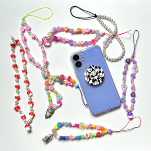 Op de afbeelding: Een verzameling kleurrijke kralen telefoonbandjes met verschillende bedels, waaronder een panda, een kat en een melkpak. De bandjes zijn gemaakt van verschillende gekleurde kralen en zijn bevestigd aan een paarse telefoonhoes met een popsocket.