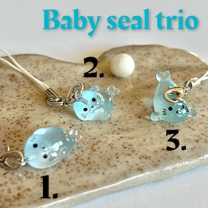 May include: Three light blue baby seal charms with black eyes and silver rings. The charms are hanging from white strings with silver clasps. The charms are numbered 1, 2, and 3 on a light brown background.