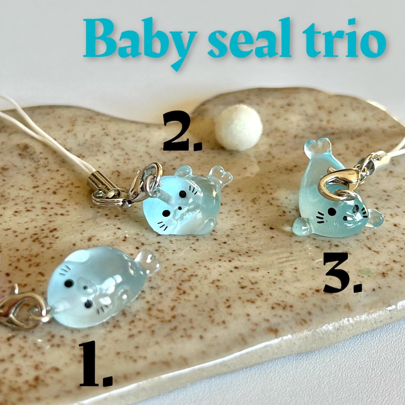 May include: Three light blue baby seal charms with black eyes and silver rings. The charms are hanging from white strings with silver clasps. The charms are numbered 1, 2, and 3 on a light brown background.