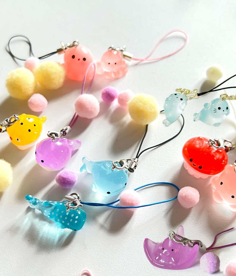 May include: A collection of colorful, handmade phone charms featuring various sea creatures, including jellyfish, octopuses, whales, and fish. The charms are made of clear resin and have a glossy finish. They are attached to colorful pom-poms and black, white, and blue cords.